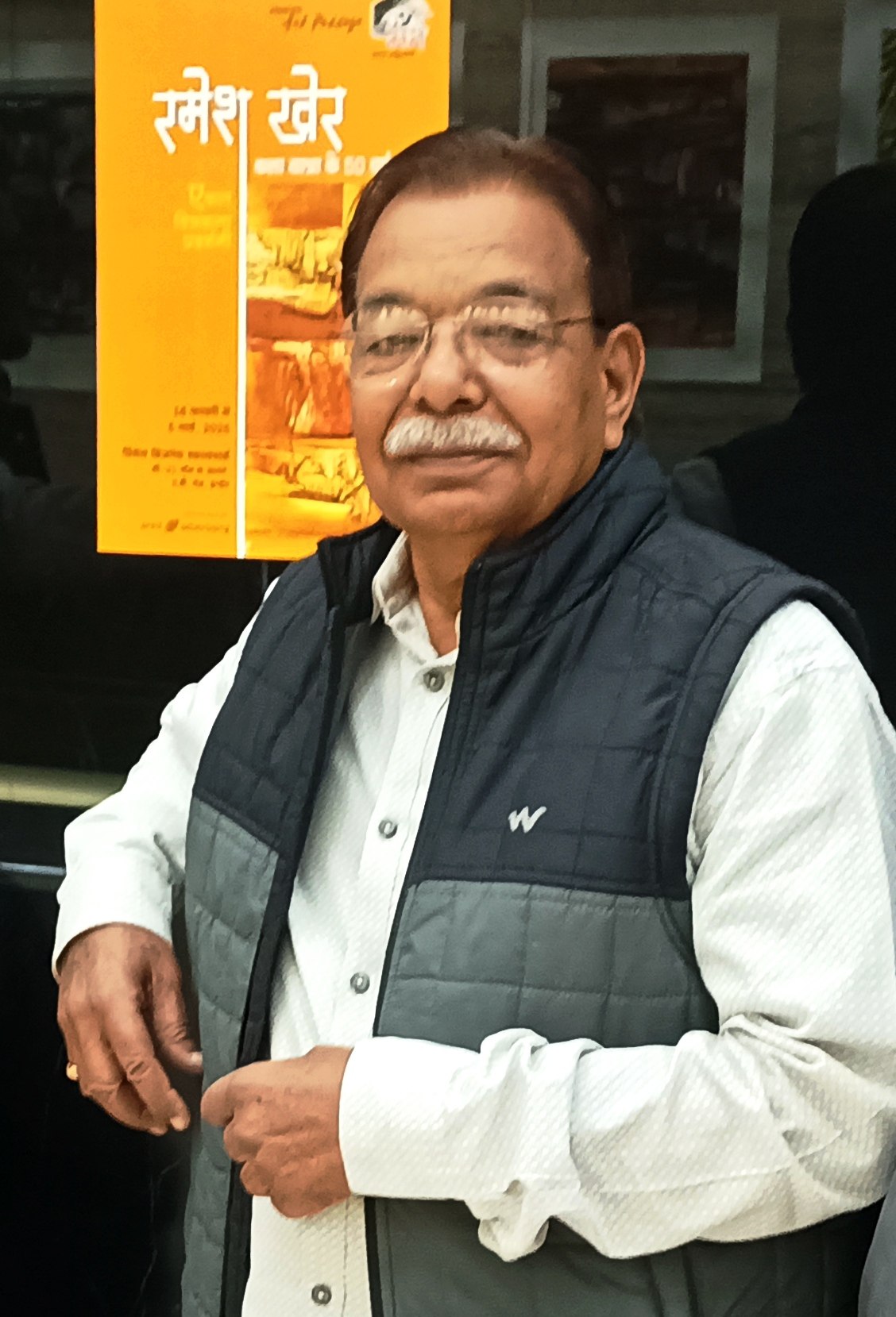 Indian abstract painter Ramesh Kher seated beside an exhibition poster bearing his name